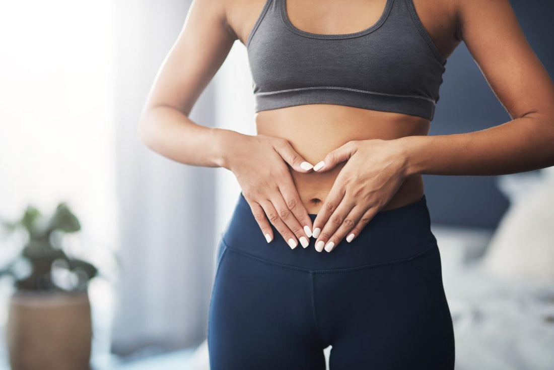 Woman in grey workout clothes standing in her bedroom with her hands placed gently over her belly button.