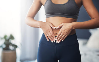 Woman in grey workout clothes standing in her bedroom with her hands placed gently over her belly button.