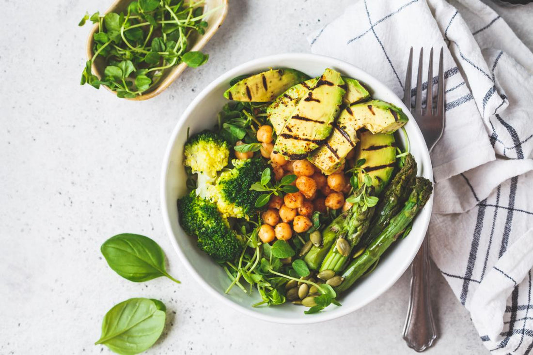 Keto-friendly dinner bowl filled with healthy foods like broccoli, asparagus, and avocado sitting on a white tabletop next to a checkered napkin and fork. 
