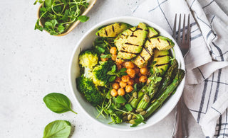 Keto-friendly dinner bowl filled with healthy foods like broccoli, asparagus, and avocado sitting on a white tabletop next to a checkered napkin and fork. 