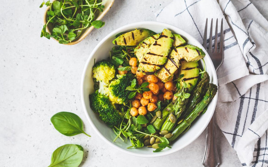 Keto-friendly dinner bowl filled with healthy foods like broccoli, asparagus, and avocado sitting on a white tabletop next to a checkered napkin and fork. 