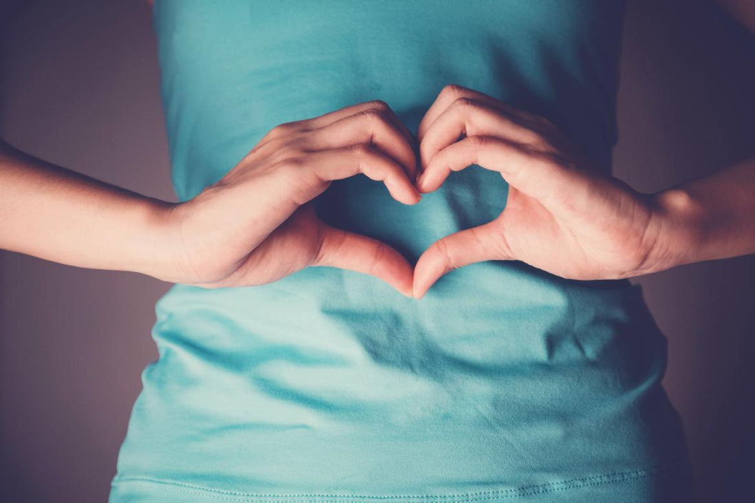 Female wearing a blue shirt holding her hands in a heart shape over her midsection. 