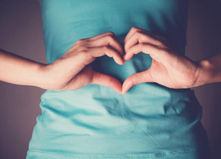 Female wearing a blue shirt holding her hands in a heart shape over her midsection. 