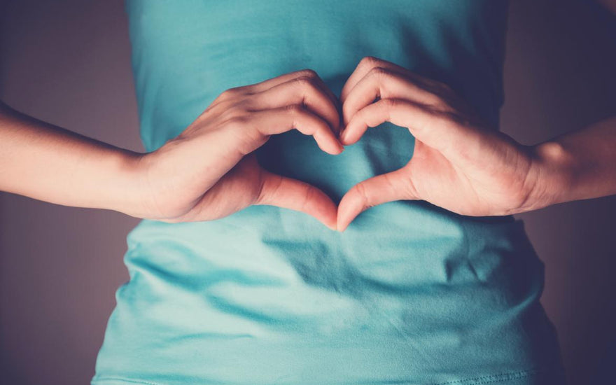 Female wearing a blue shirt holding her hands in a heart shape over her midsection. 