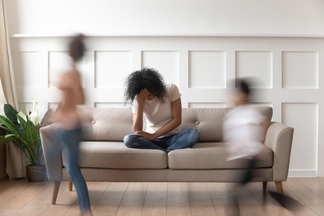 Stressed out mother sitting on the couch with her head in her hands while her kids run around in front of her.