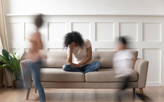 Stressed out mother sitting on the couch with her head in her hands while her kids run around in front of her.