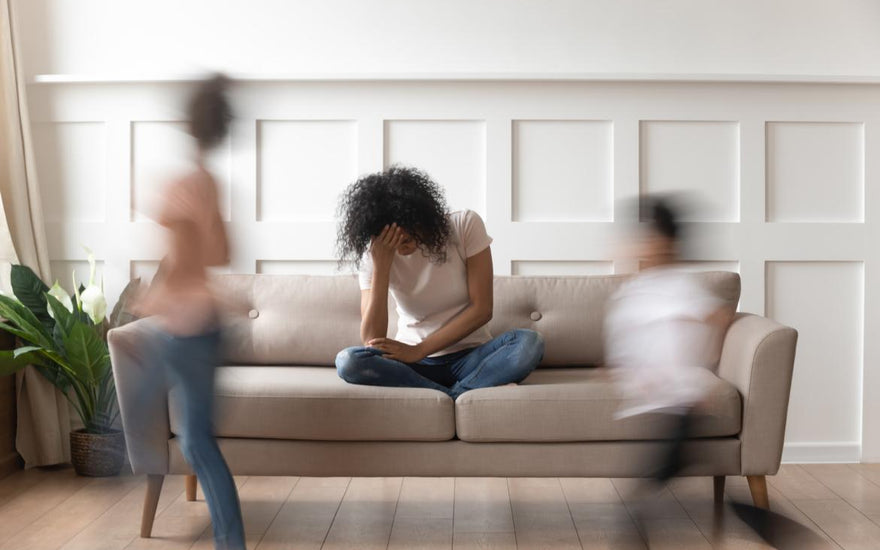 Stressed out mother sitting on the couch with her head in her hands while her kids run around in front of her.