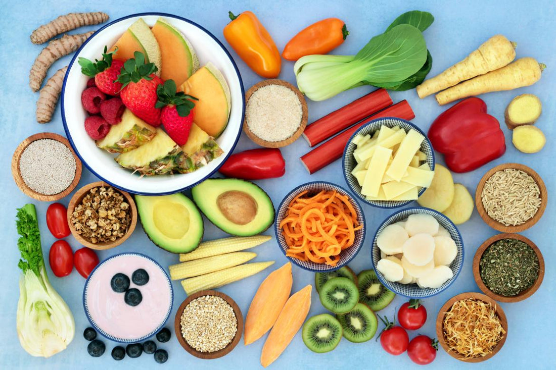Flat lay of a variety of healthy whole foods, including avocados, kiwis, berries, peppers, seeds, and nuts, on a light blue background.