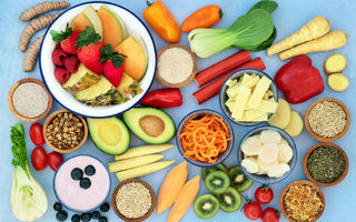 Flat lay of a variety of healthy whole foods, including avocados, kiwis, berries, peppers, seeds, and nuts, on a light blue background.