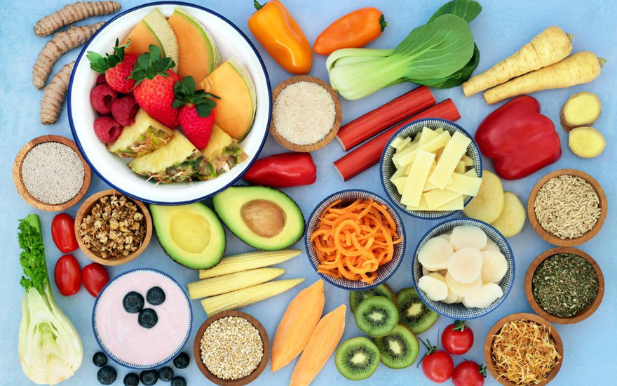 Flat lay of a variety of healthy whole foods, including avocados, kiwis, berries, peppers, seeds, and nuts, on a light blue background.