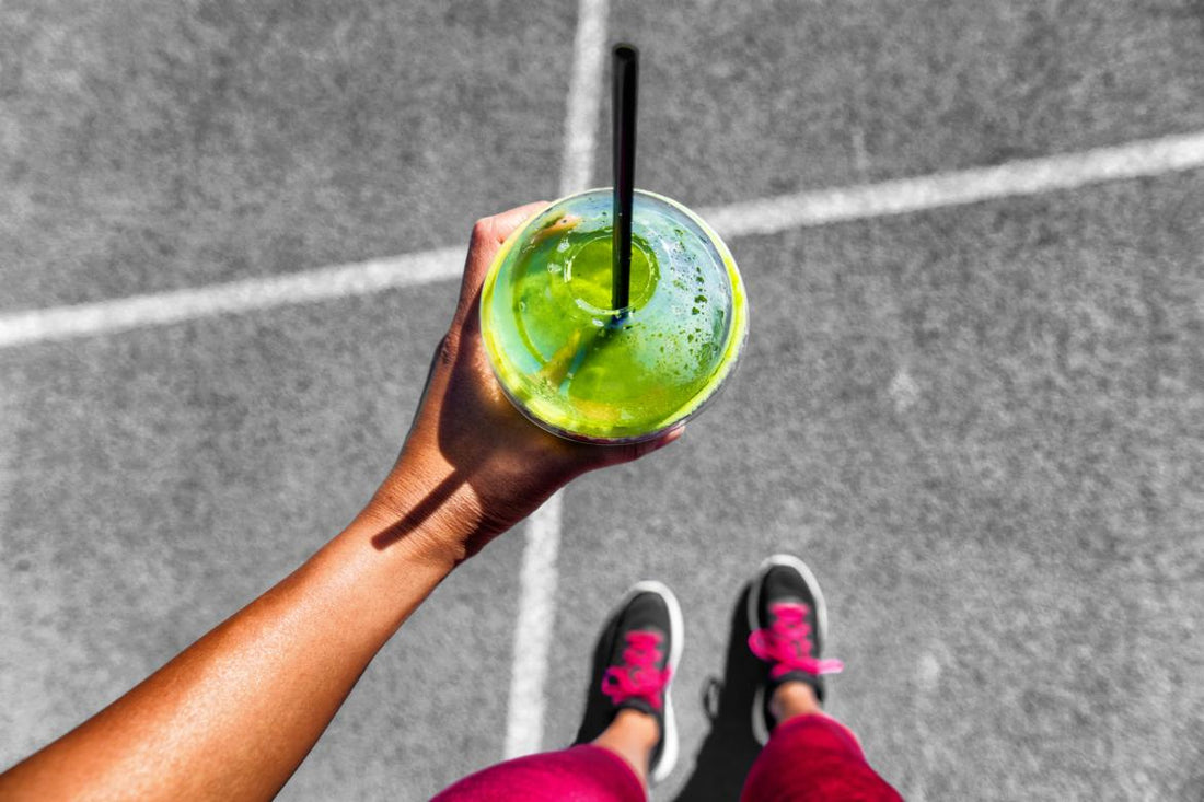Top down view of a green smoothie in a to-go cup being held by a woman in running shoes.