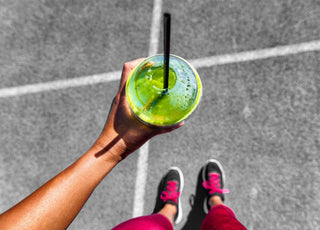 Top down view of a green smoothie in a to-go cup being held by a woman in running shoes.