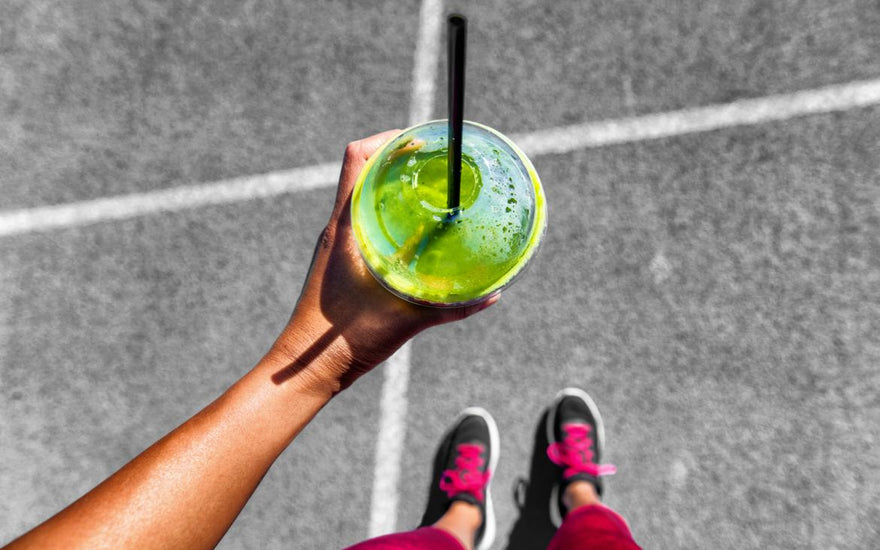 Top down view of a green smoothie in a to-go cup being held by a woman in running shoes.