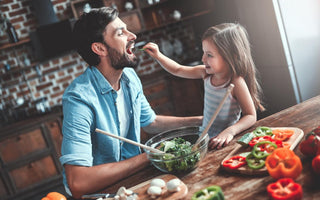 Young girl feeding her dad a piece of a green vegetable at the dinner table. 