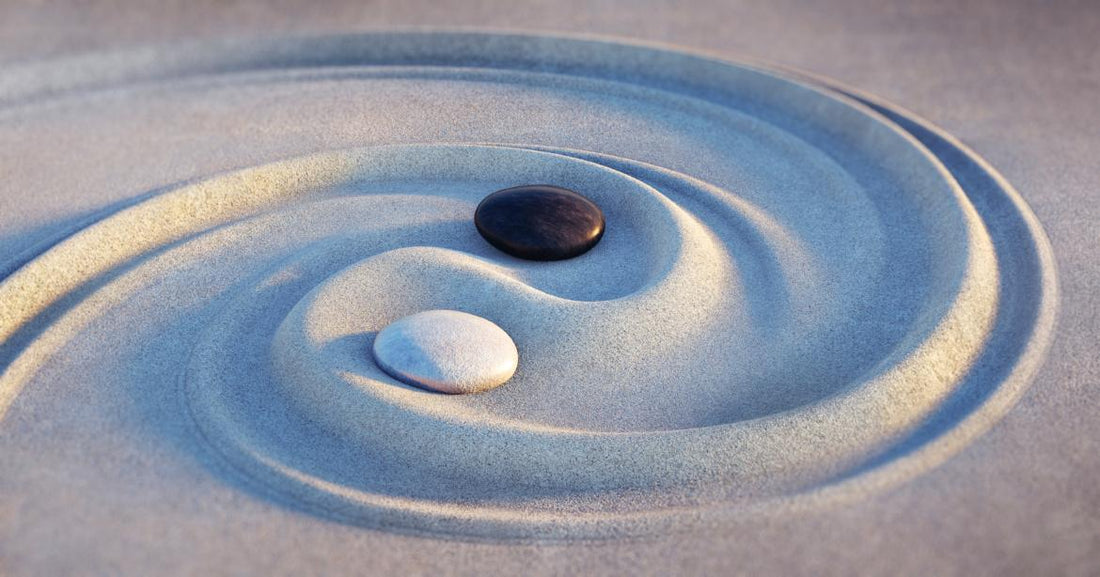 Two pebbles, one black and one white, sitting in the middle of a swirl drawn in the sand. 