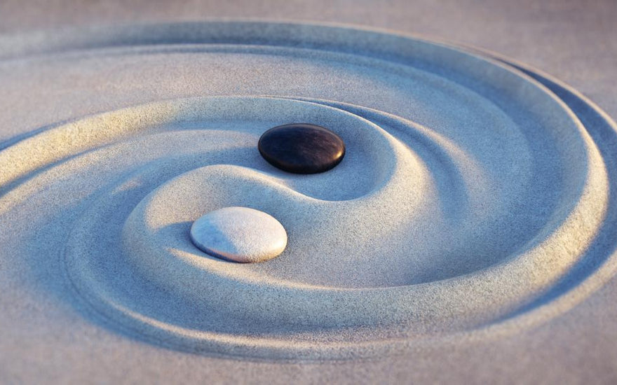 Two pebbles, one black and one white, sitting in the middle of a swirl drawn in the sand. 