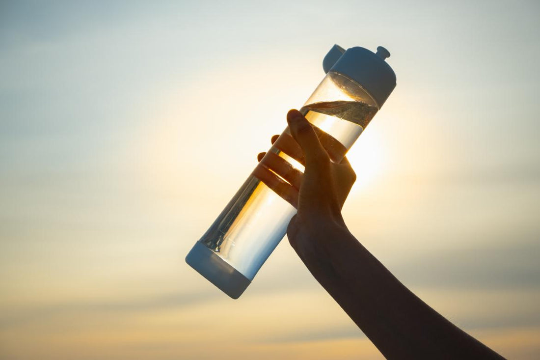 Female hand holding a clear water bottle full of water up in the air at sunset. 