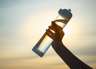 Female hand holding a clear water bottle full of water up in the air at sunset. 