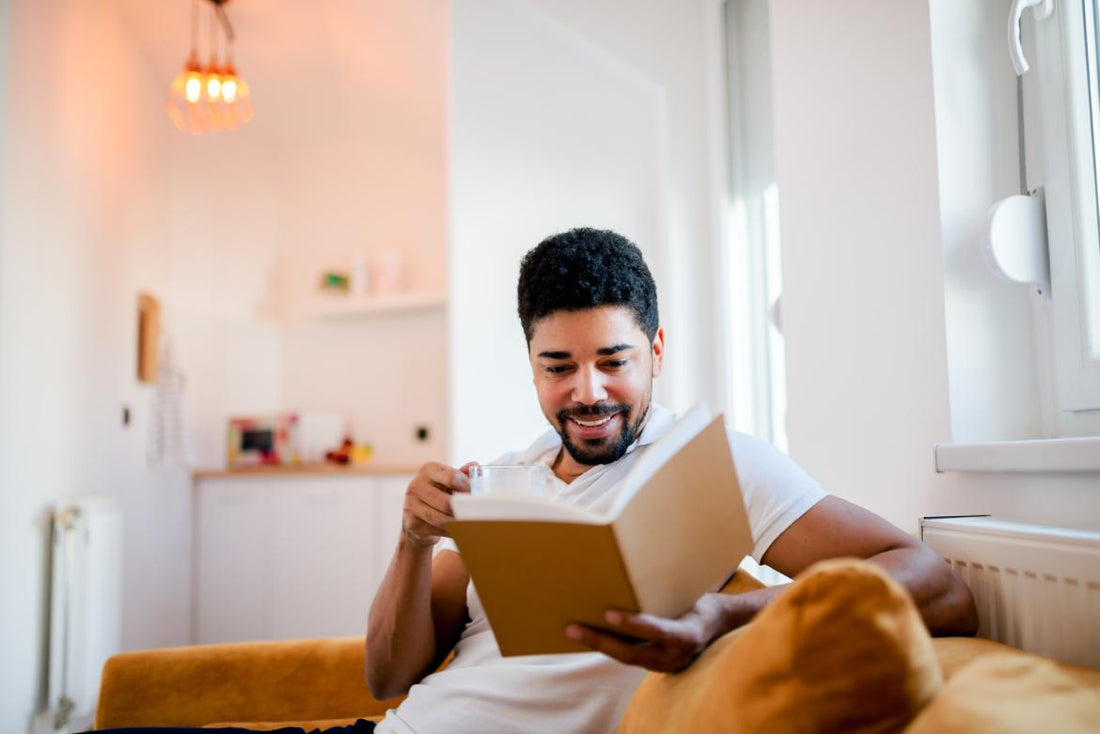 Adult male sittin gon the couch in a living room with a glass of water while reading a book.