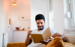 Adult male sittin gon the couch in a living room with a glass of water while reading a book.