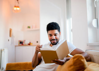 Adult male sittin gon the couch in a living room with a glass of water while reading a book.