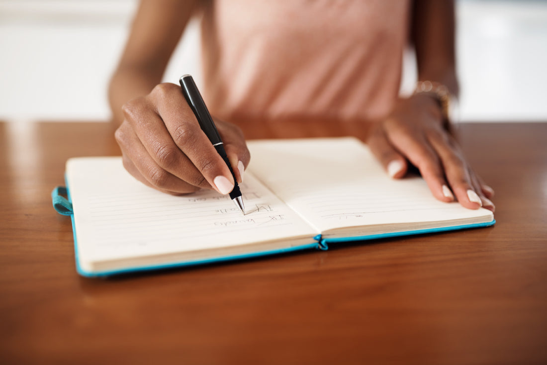 Woman in a pink shirt sitting at a wooden table writing in a blue journal.