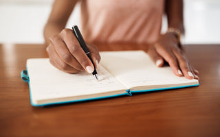 Woman in a pink shirt sitting at a wooden table writing in a blue journal.