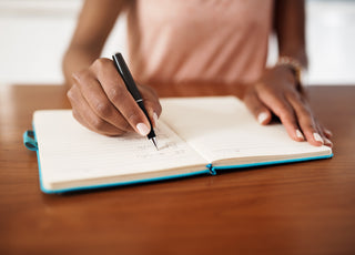 Woman in a pink shirt sitting at a wooden table writing in a blue journal.