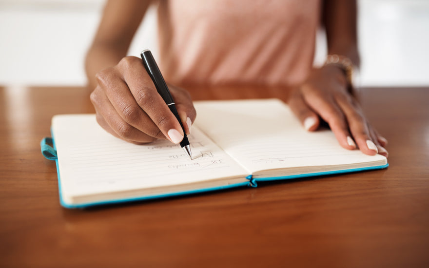 Woman in a pink shirt sitting at a wooden table writing in a blue journal.