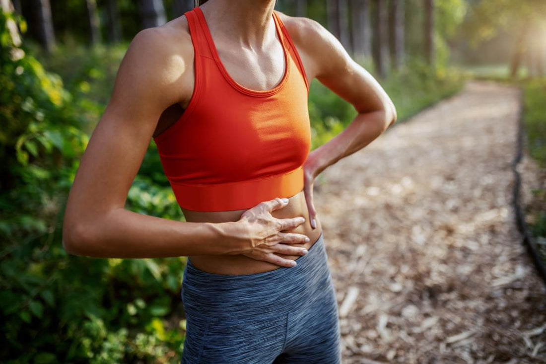 Female in orange and grey workout gear standing on an outdoor running trail with a hand on her stomach.