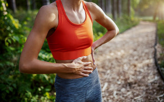 Female in orange and grey workout gear standing on an outdoor running trail with a hand on her stomach.