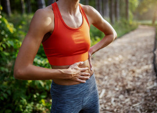 Female in orange and grey workout gear standing on an outdoor running trail with a hand on her stomach.