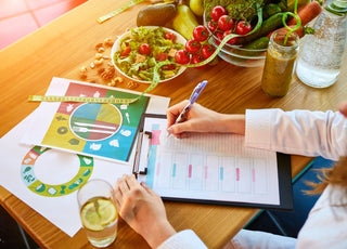 Woman sitting at a table with different healthy ingredients across it while she writes an eating plan.