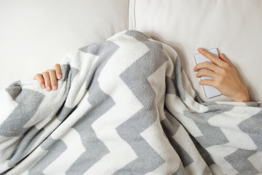 Person laying in bed with a grey and white striped blanket pulled over their head so you can only see their hands.