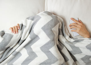 Person laying in bed with a grey and white striped blanket pulled over their head so you can only see their hands.