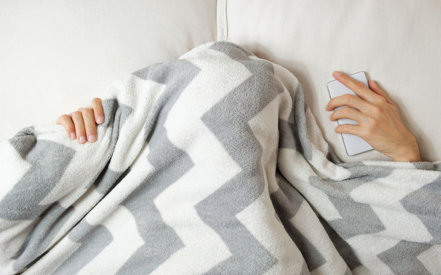 Person laying in bed with a grey and white striped blanket pulled over their head so you can only see their hands.