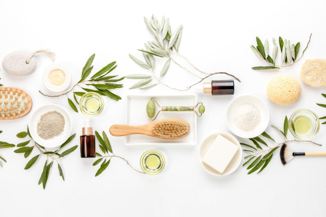 Flat lay of skin care items such as cleansers, oils, and pumice stones on a table decorated with greenery.