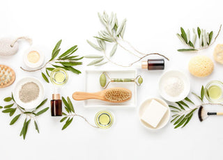 Flat lay of skin care items such as cleansers, oils, and pumice stones on a table decorated with greenery.