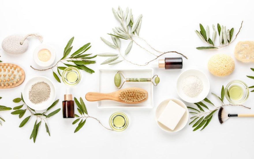 Flat lay of skin care items such as cleansers, oils, and pumice stones on a table decorated with greenery.