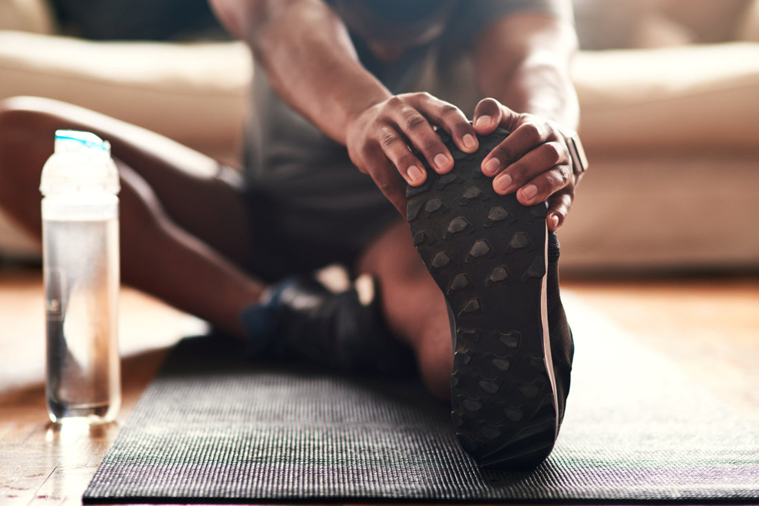 Man sitting on a black yoga matt with one leg out in front of him as he stretches.