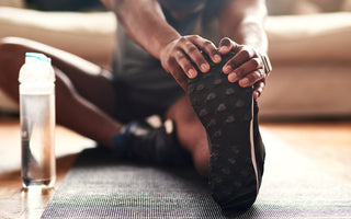 Man sitting on a black yoga matt with one leg out in front of him as he stretches.