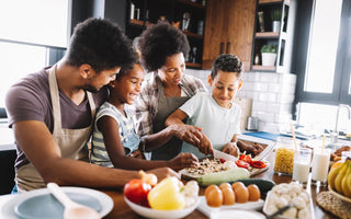 Family of four grouped around a cutting board with the youngest child chopping mushrooms while they cook dinner together.