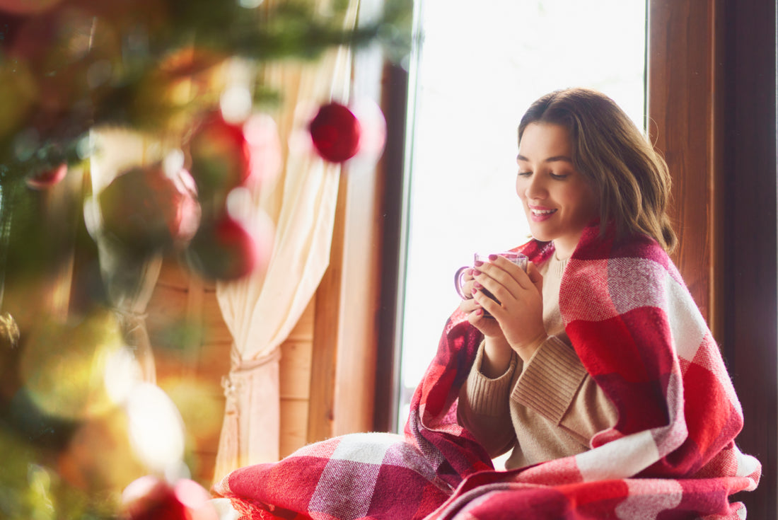 Young adult woman sitting in front of a Christmas tree with a red checkered blanket wrapped around her and a warm beverage in her hands.