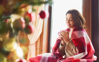 Young adult woman sitting in front of a Christmas tree with a red checkered blanket wrapped around her and a warm beverage in her hands.