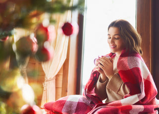 Young adult woman sitting in front of a Christmas tree with a red checkered blanket wrapped around her and a warm beverage in her hands.