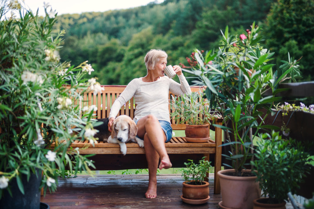 Older woman with short grey hair in jean shorts and white shirt sitting outside on a wooden bench sipping her coffee and petting her dog that is sleeping on the bench next to her. 