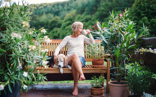 Older woman with short grey hair in jean shorts and white shirt sitting outside on a wooden bench sipping her coffee and petting her dog that is sleeping on the bench next to her. 