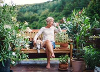 Older woman with short grey hair in jean shorts and white shirt sitting outside on a wooden bench sipping her coffee and petting her dog that is sleeping on the bench next to her. 