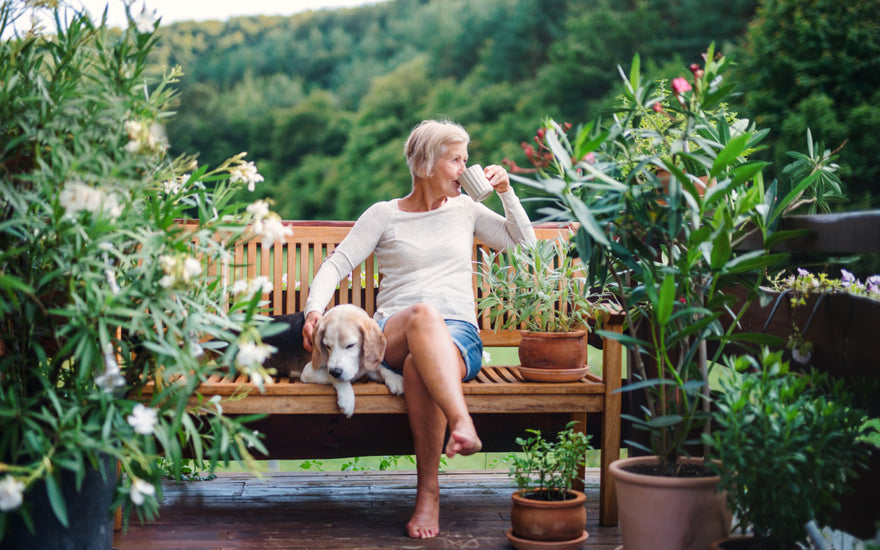 Older woman with short grey hair in jean shorts and white shirt sitting outside on a wooden bench sipping her coffee and petting her dog that is sleeping on the bench next to her. 