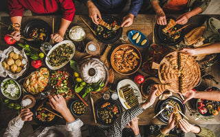 Birds-eye view of a wooden table full of common holiday foods as people reach toward items to serve themselves.